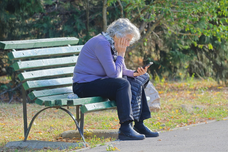 11 - An elderly woman with short, grey-white hair is seated on a green slatted wooden park bench, wearing a purple sweater, dark pants, black shoes, and a patterned scarf, while looking down at a mobile phone